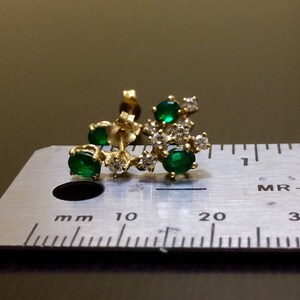 May include: A gold earring with a curved design featuring five green gemstones and small clear gemstones. The earring is measured against a ruler.