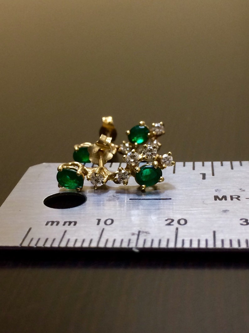 May include: A gold earring with a curved design featuring five green gemstones and small clear gemstones. The earring is measured against a ruler.