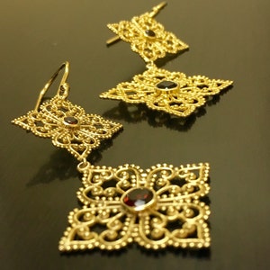 May include: A pair of gold filigree earrings with dangling square-shaped charms. Each charm features a red gemstone in the centre.