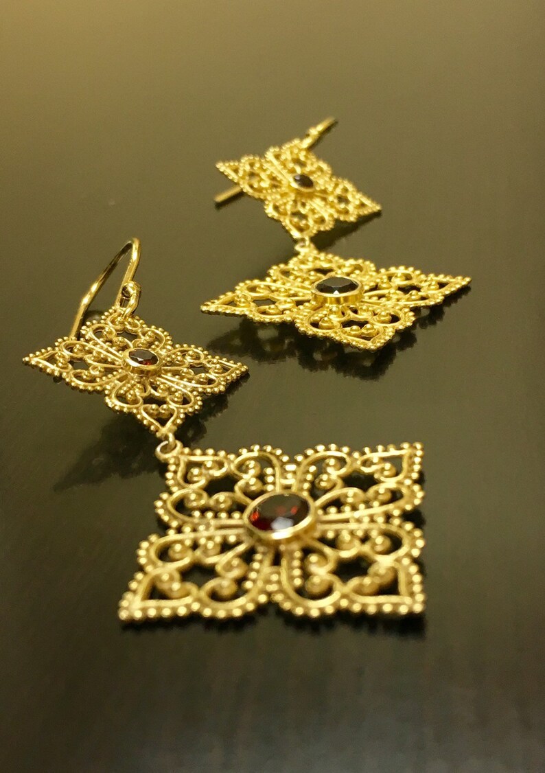 May include: A pair of gold filigree earrings with dangling square-shaped charms. Each charm features a red gemstone in the centre.