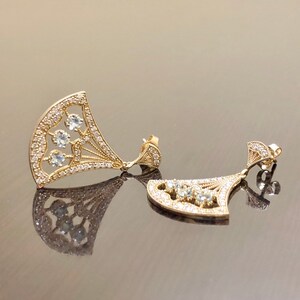 May include: A pair of gold earrings with a fan-shaped design and clear stones. The earrings are dangling and have a delicate, intricate design.