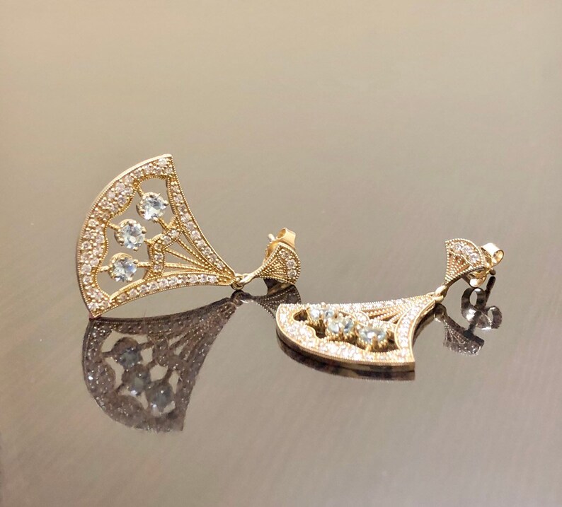 May include: A pair of gold earrings with a fan-shaped design and clear stones. The earrings are dangling and have a delicate, intricate design.