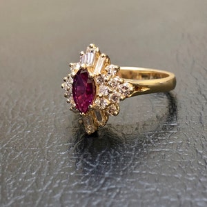 May include: A gold ring featuring a large, oval-shaped ruby surrounded by smaller diamonds. The ruby is a deep red colour and the diamonds are clear.