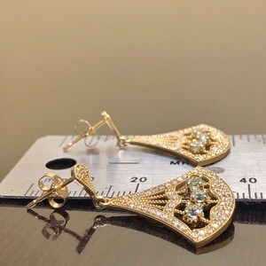 May include: A pair of gold dangle earrings with a teardrop shape. The earrings are adorned with small, clear stones and larger, clear stones in the center. The earrings are shown on a ruler, measuring approximately 1.5 inches in length.