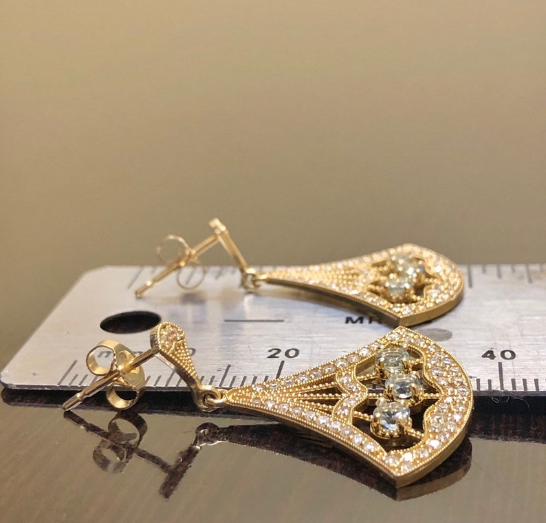 May include: A pair of gold dangle earrings with a teardrop shape. The earrings are adorned with small, clear stones and larger, clear stones in the center. The earrings are shown on a ruler, measuring approximately 1.5 inches in length.