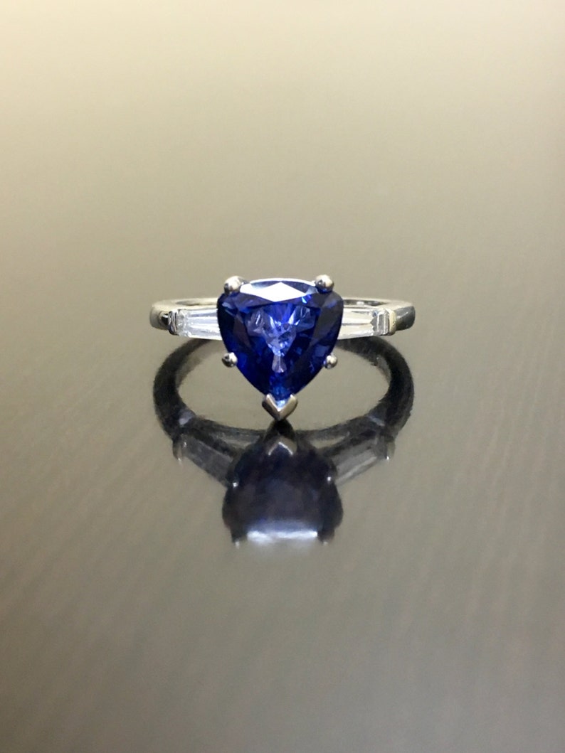 May include: A heart-shaped blue sapphire engagement ring set in a silver band with two smaller diamonds on either side of the sapphire.