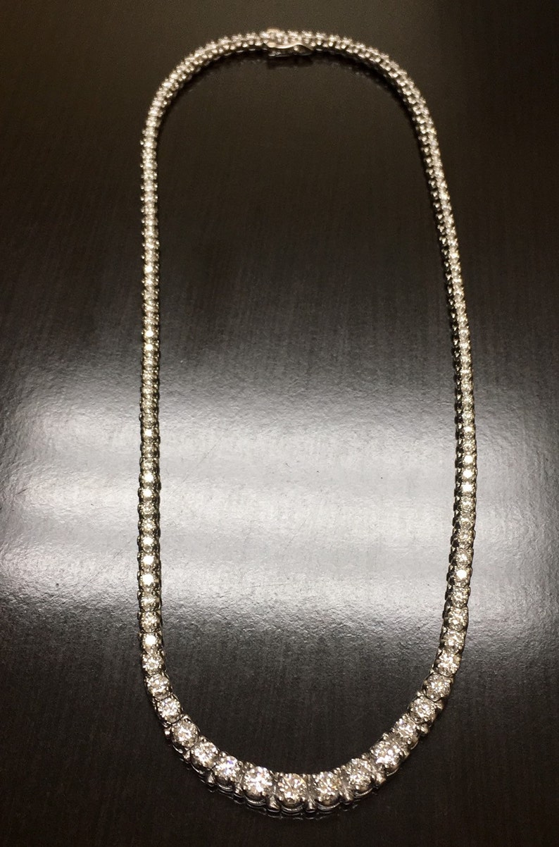 May include: A silver necklace with a row of round, clear stones. The necklace is on a dark background.