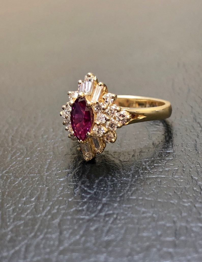 Op de afbeelding: Een gouden ring met een grote, ovale robijn omringd door kleinere diamanten. De robijn is diep rood van kleur en de diamanten zijn helder.