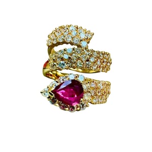 May include: A gold-toned ring with a spiral design, encrusted with numerous small, clear gemstones. The ring features a large, pear-shaped, deep-red gemstone at the base. The ring is set against a white background.