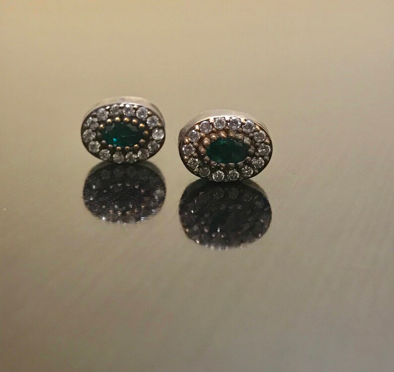 May include: A pair of silver oval earrings with a green gemstone surrounded by a halo of smaller clear gemstones.