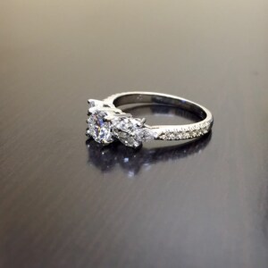 May include: A silver ring featuring a large, round, clear centre stone flanked by two smaller, pear-shaped stones. The band is set with smaller, clear stones.