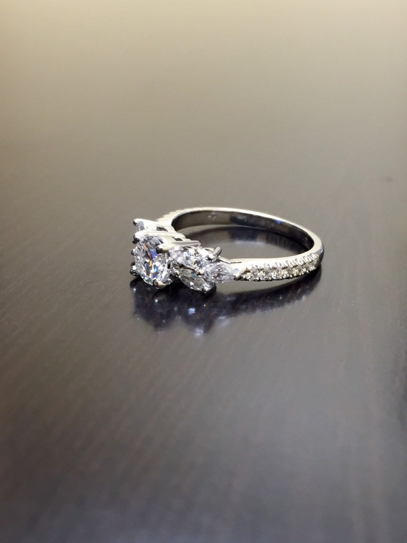 May include: A silver ring featuring a large, round, clear centre stone flanked by two smaller, pear-shaped stones. The band is set with smaller, clear stones.