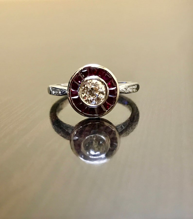 May include: A silver ring with a round, bezel-set diamond surrounded by a halo of red rubies.