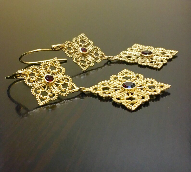 May include: Gold filigree earrings with red gemstone accents. The earrings feature a delicate, intricate design with a dangling drop.
