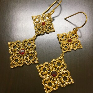 May include: A pair of gold filigree earrings with red gemstone accents. The earrings feature a delicate, intricate design with multiple layers of filigree.