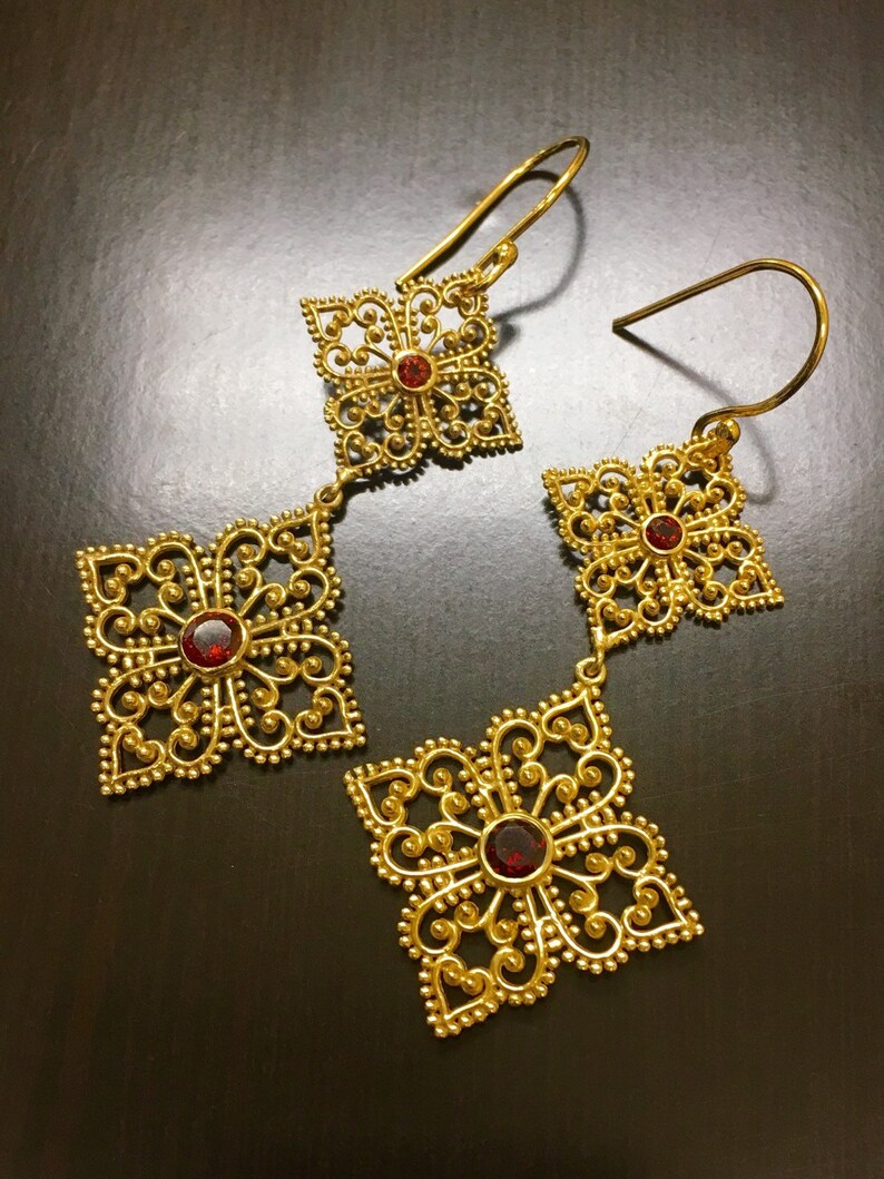 May include: A pair of gold filigree earrings with red gemstone accents. The earrings feature a delicate, intricate design with multiple layers of filigree.