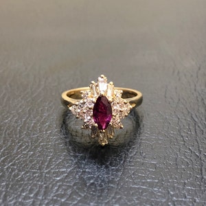 May include: A gold ring featuring a large, marquise-shaped ruby surrounded by smaller diamonds. The ring has a delicate, floral design.