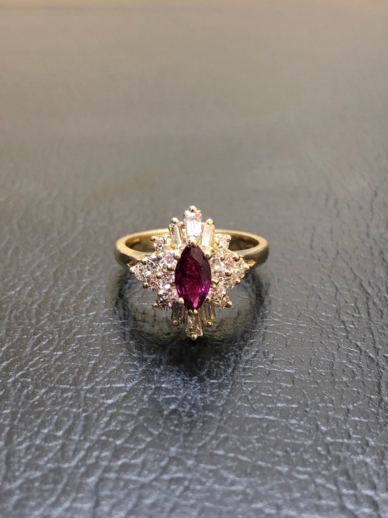 May include: A gold ring featuring a large, marquise-shaped ruby surrounded by smaller diamonds. The ring has a delicate, floral design.