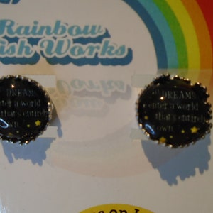 May include: Two silver-toned round earrings with black backgrounds and white text that reads "DREAMS enter a world that's entirely your own". The earrings have a silver-toned border and are decorated with small yellow stars.