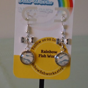 May include: Silver-toned dangle earrings. Each earring features a small bow-shaped charm above a round pendant with a blue and white abstract design. The earrings hang from a silver hook. The card behind the earrings says "Rainbow Fish Works".