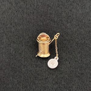 May include: A gold-toned metal sewing spool charm featuring a ribbed cylindrical body and a thin gold chain attached to a small, round, white button-shaped pendant with a four-hole design, presented as a detailed miniature sewing accessory.