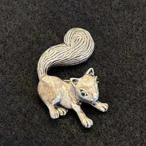 May include: A silver-toned metal brooch shaped like a squirrel with a textured, curled tail and small green inset stones for eyes. The squirrel is depicted in a pouncing or playing position with its front paws extended forward and head turned.