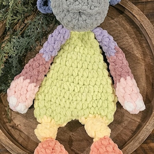 Puede incluir: Muñeca de ganchillo hecha a mano con cabeza gris, orejas azules y ojos negros. La muñeca tiene un cuerpo verde claro, brazos morados y rosas y piernas color melocotón. Acentos blancos en los pies. La muñeca está sobre una bandeja de madera.