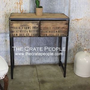Puede incluir: Una mesa consola de madera con dos cajones hechos de cajas de madera reutilizadas. Los cajones tienen letras negras que dicen "Zoria Farms Inc. Brentwood & Madera San Jose CA 1994". La mesa tiene un marco de metal negro y se encuentra sobre un suelo de hormigón.