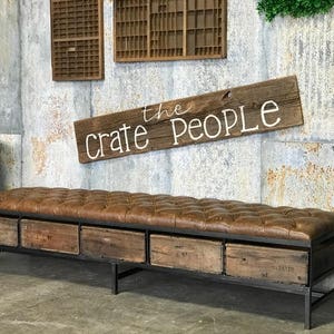 May include: A brown leather tufted bench with a metal frame and six wooden drawers. The bench is in front of a wall with a wooden sign that says "the crate people".