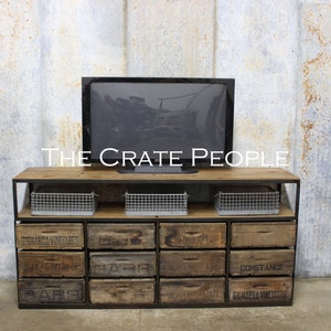 May include: A rustic wooden TV stand with metal framing and drawers. The drawers are made from repurposed wooden crates with labels like "Constande" and "Guadalajara Vinedares". The stand has three wire baskets on the top shelf. The stand is in front of a gray wall with a flat screen TV on top.