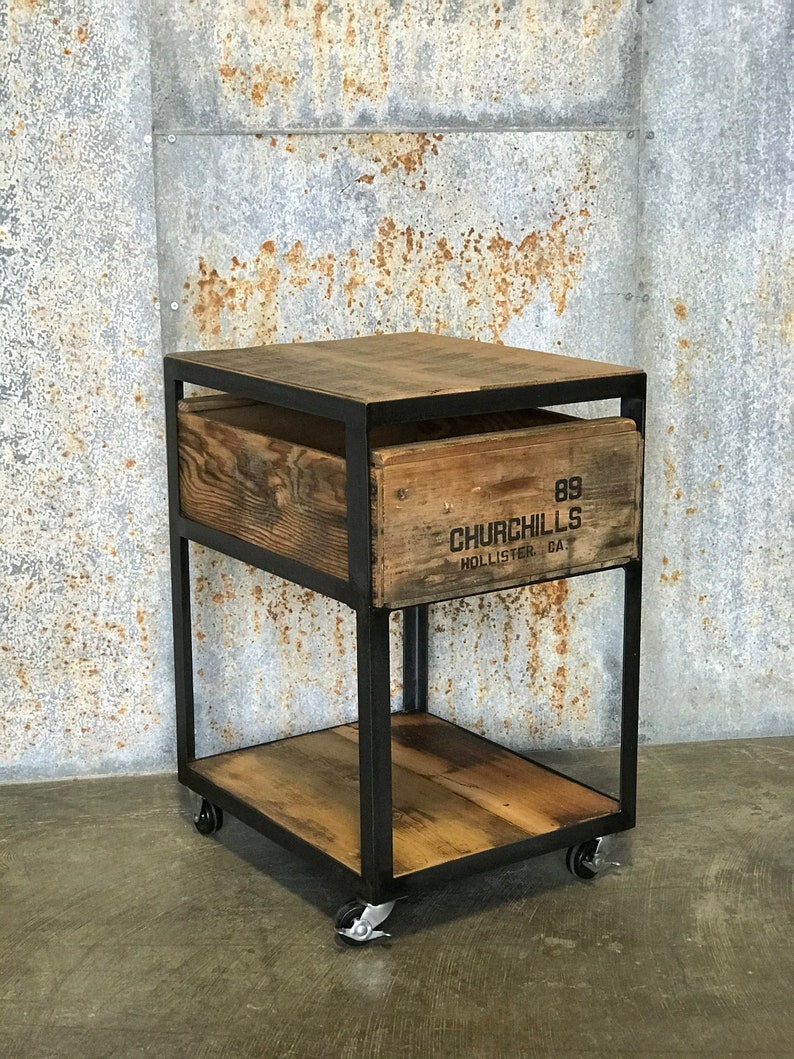 Puede incluir: Un carro rodante de metal negro con dos estantes de madera. El estante superior est&aacute; hecho de una caja de madera reutilizada con el texto "89 CHURCHILLS HOLLISTER, GA." impreso en el lateral.