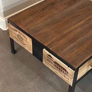 Puede incluir: Una mesa de café de madera con un marco de metal negro. La superficie de la mesa está hecha de madera recuperada y tiene un acabado natural. La mesa tiene dos cajones, uno a cada lado. Los cajones están hechos de madera y tienen etiquetas que dicen "Cap Royal" y "Chateau Talbot".