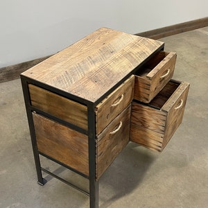 May include: A wooden desk with three drawers, each with rope handles. The desk is made of reclaimed wood and has a rustic look. The desk is on a concrete floor.
