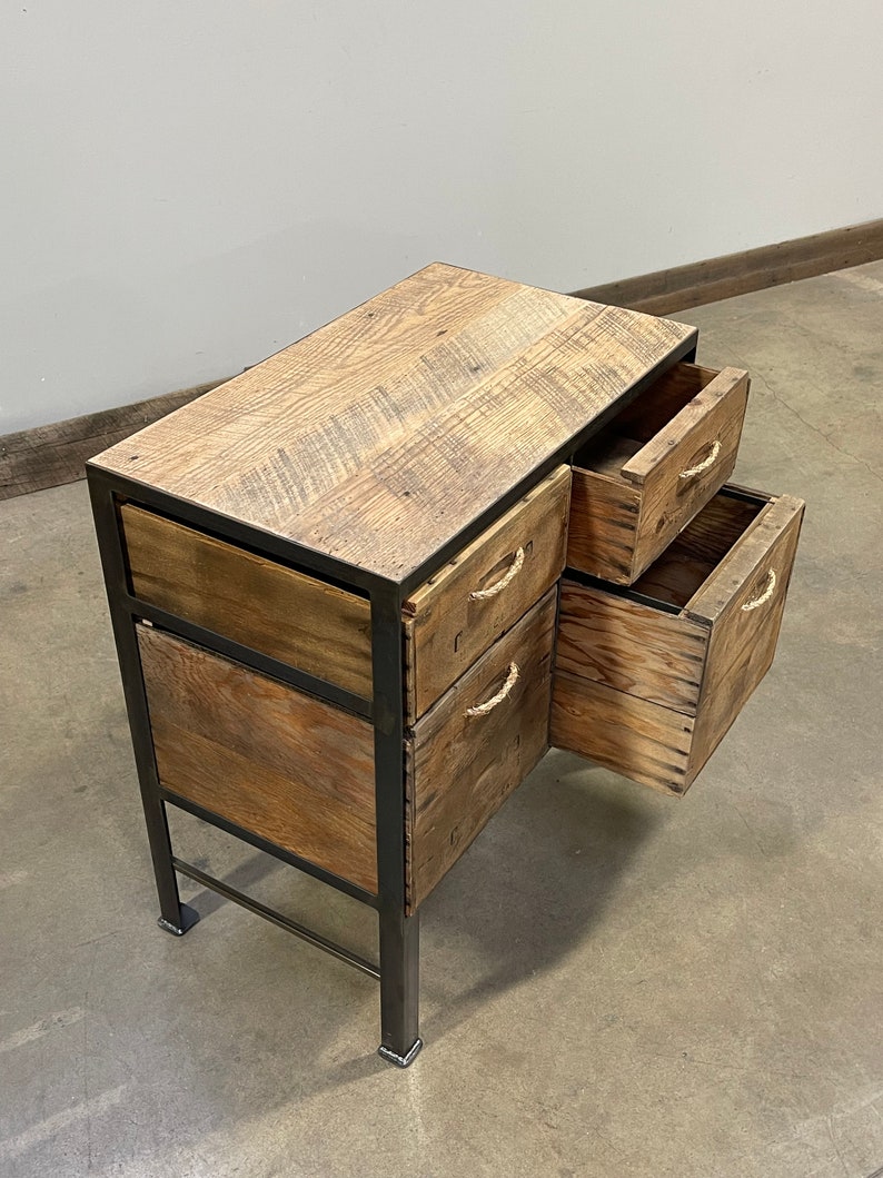 May include: A wooden desk with three drawers, each with rope handles. The desk is made of reclaimed wood and has a rustic look. The desk is on a concrete floor.