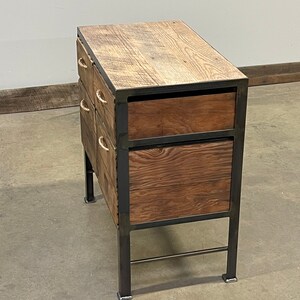 May include: A wooden chest of drawers with a metal frame and rope handles. The drawers are stacked in a staggered pattern, with the top drawer being the largest. The chest is sitting on a concrete floor.