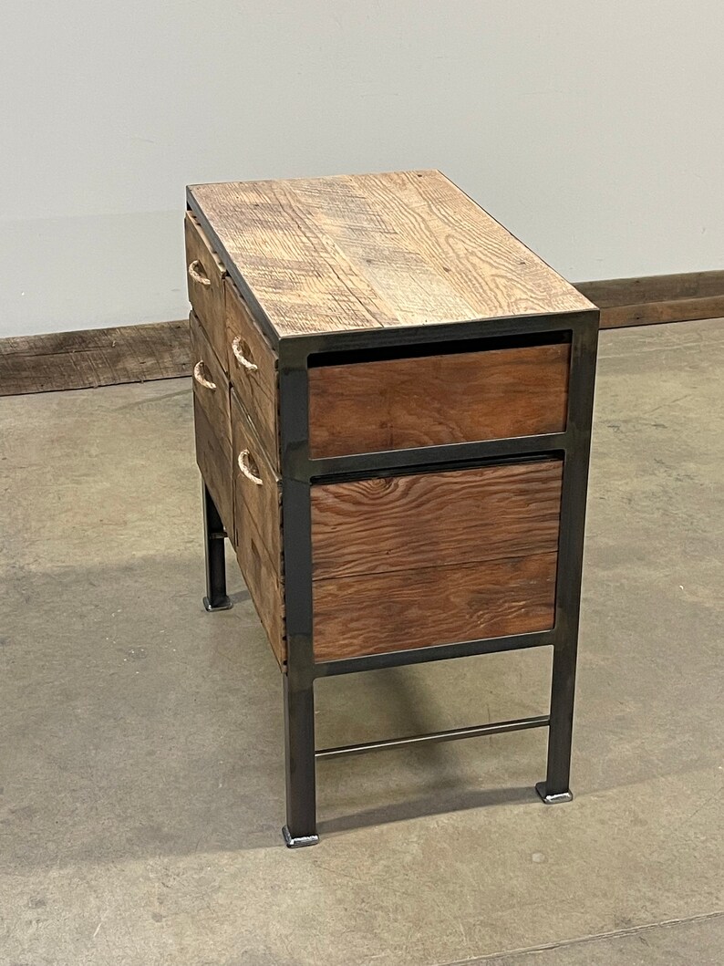 May include: A wooden chest of drawers with a metal frame and rope handles. The drawers are stacked in a staggered pattern, with the top drawer being the largest. The chest is sitting on a concrete floor.