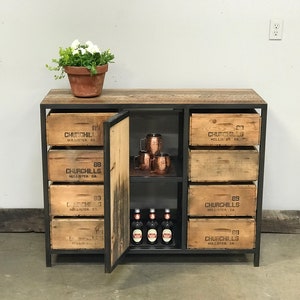 Puede incluir: Un armario de metal negro con seis cajones y una puerta. Los cajones son de madera y tienen el texto "Churchills Hollister CA" impreso en ellos. El armario tiene un estante dentro de la puerta con tazas de cobre y botellas.