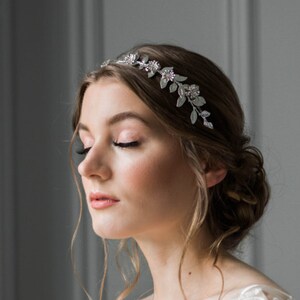 May include: A woman wearing a white floral lace dress with a silver leaf and crystal headband. The dress has a V-neckline and short sleeves.