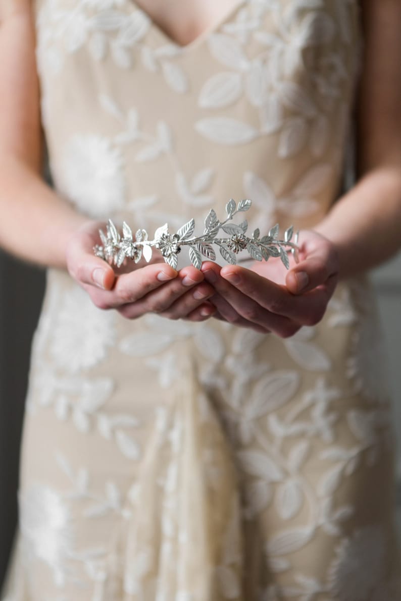 May include: A silver floral crown with delicate leaves and small flowers. The crown is held in the hands of a person wearing a beige dress with white floral embroidery.