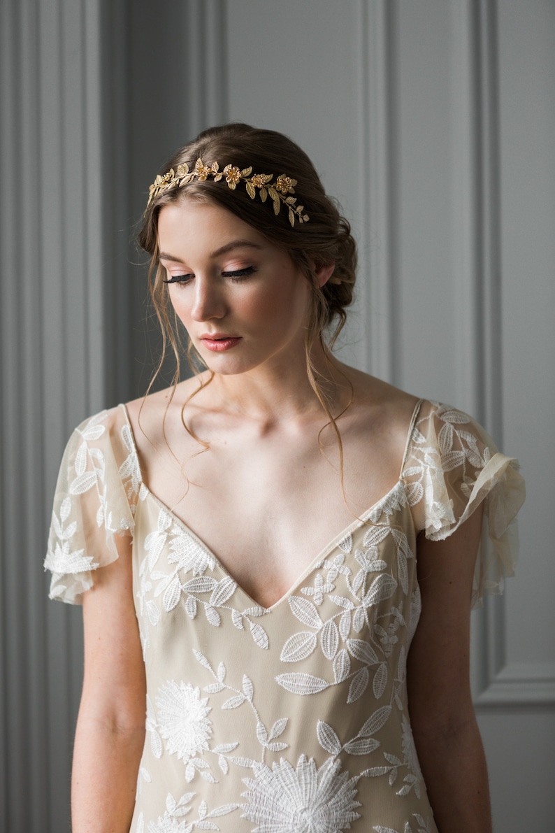 May include: A woman wearing a beige dress with white floral embroidery and a gold leaf and flower crown.