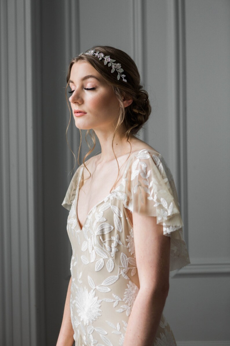 May include: A woman wearing a white floral lace dress with a silver leaf and crystal headband. The dress has a V-neckline and short sleeves.