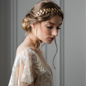 May include: A woman wearing a gold leaf crown headband, with her hair styled in a loose bun, is wearing a beige lace dress with a floral pattern.