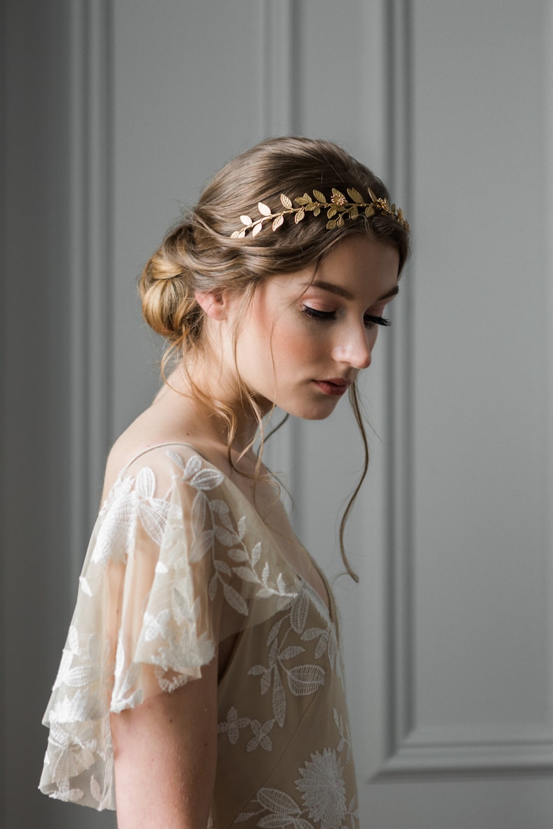 May include: A woman wearing a gold leaf crown headband, with her hair styled in a loose bun, is wearing a beige lace dress with a floral pattern.