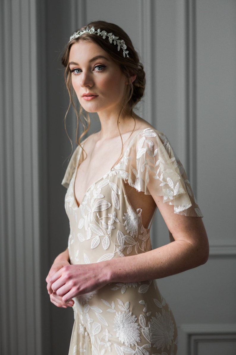May include: A woman wearing a white floral lace dress with a delicate silver headband. The dress has a V-neckline and short sleeves.