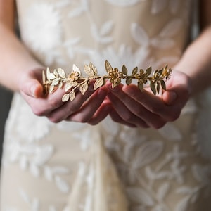 May include: A gold leaf and flower crown with delicate details. The crown is held in the hands of a person.