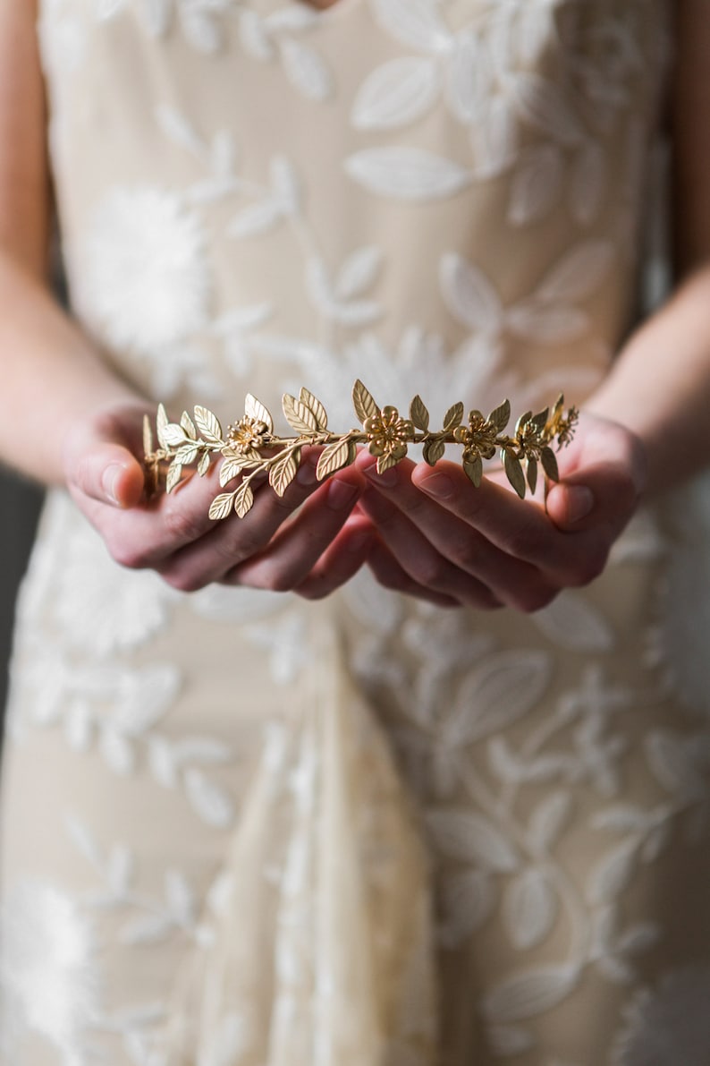 May include: A gold leaf and flower crown with delicate details. The crown is held in the hands of a person.