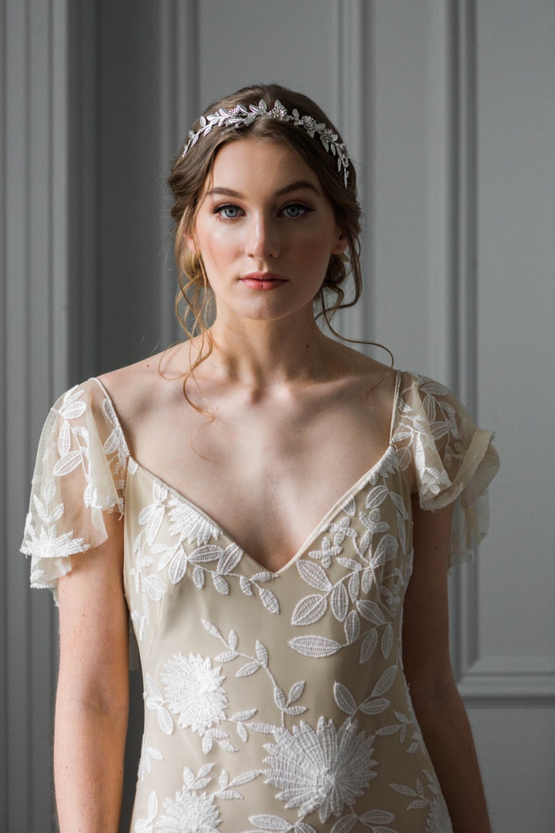 May include: A woman wearing a beige lace dress with a floral pattern and a silver leaf crown.