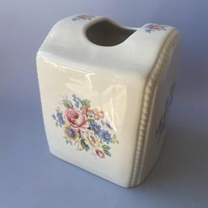 Op de afbeelding: Een crèmekleurige keramische tissueboxhouder met een rechthoekige vorm en een afgeronde opening aan de bovenkant. De buitenkant is voorzien van een kleurrijke bloemenprint met roze, gele en blauwe bloemen op de voorzijde en kleine bloemaccenten.