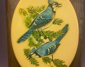 May include: Vintage oval wall art featuring two blue jays perched on green branches against a yellow background. The birds have blue and white feathers. The artwork is set in a brown wooden frame with a brass hanger.