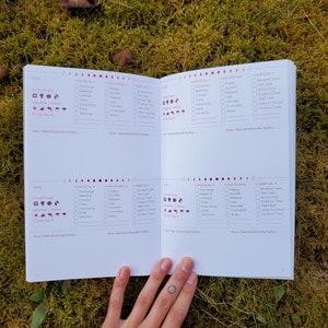May include: A white journal with a pink and purple design. The journal has a section for tracking physical, emotional, and habitual activities. The journal has a section for tracking the menstrual cycle, weather, and mood. The journal has a section for tracking the day of the cycle.